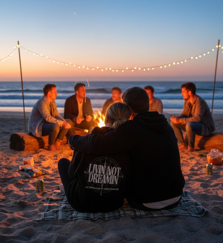 Livin’ not Dreamin’ hoodie worn by a person sitting by a beach campfire at sunset with friends