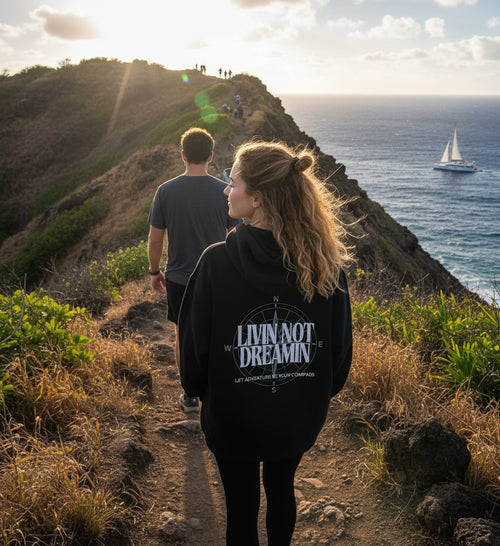 Livin’ not Dreamin’ black fleece hoodie with compass graphic worn by a woman hiking a coastal trail at sunset.