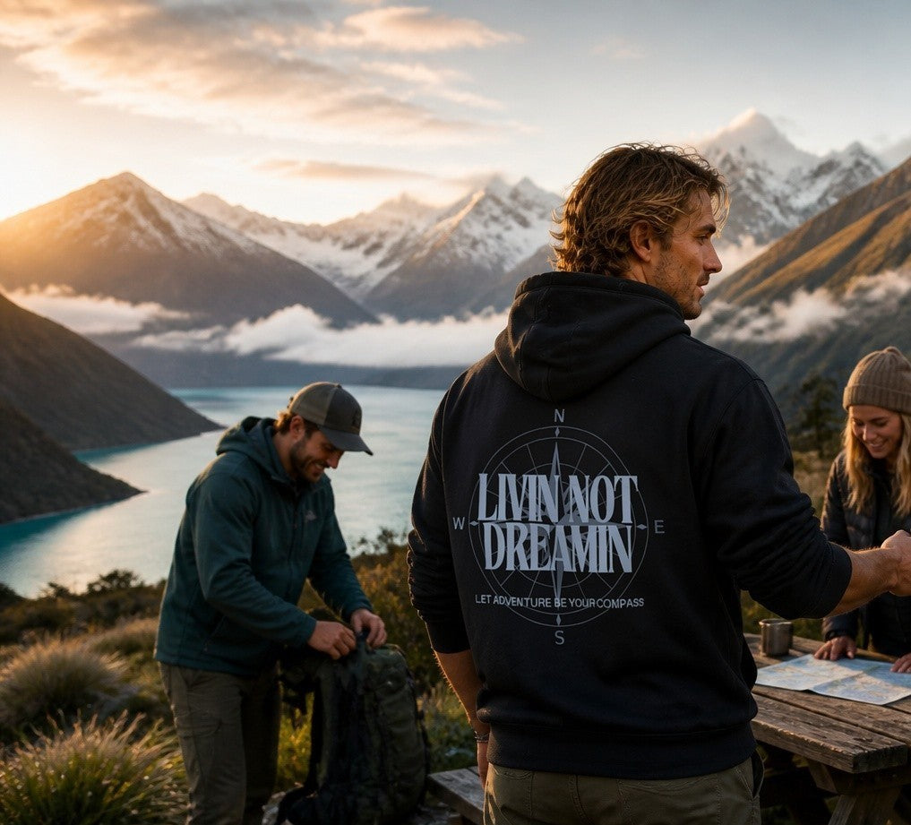 Livin’ not Dreamin’ black hoodie with compass graphic worn by a man overlooking a scenic mountain lake at sunset.
