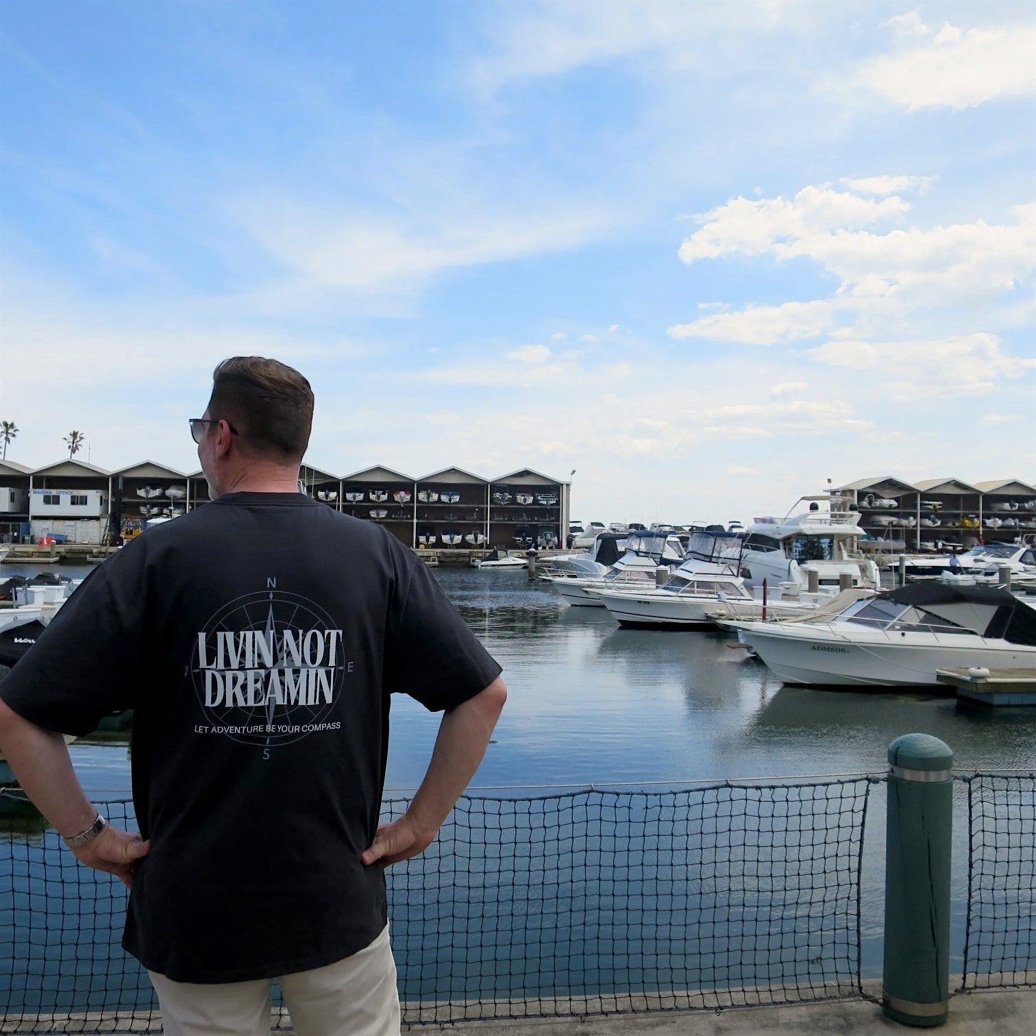 Man wearing a black Livin’ not Dreamin’ heavyweight cotton crew neck tee with a compass graphic at a marina