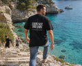Back view of a man wearing the black Livin’ not Dreamin’ Compass Tee with its white branded back compass print beside turquoise coastal water.