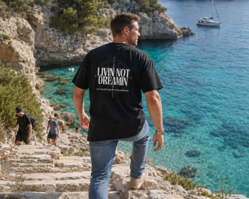 Back view of a man wearing the black Livin’ not Dreamin’ Compass Tee with its white branded back compass print beside turquoise coastal water.