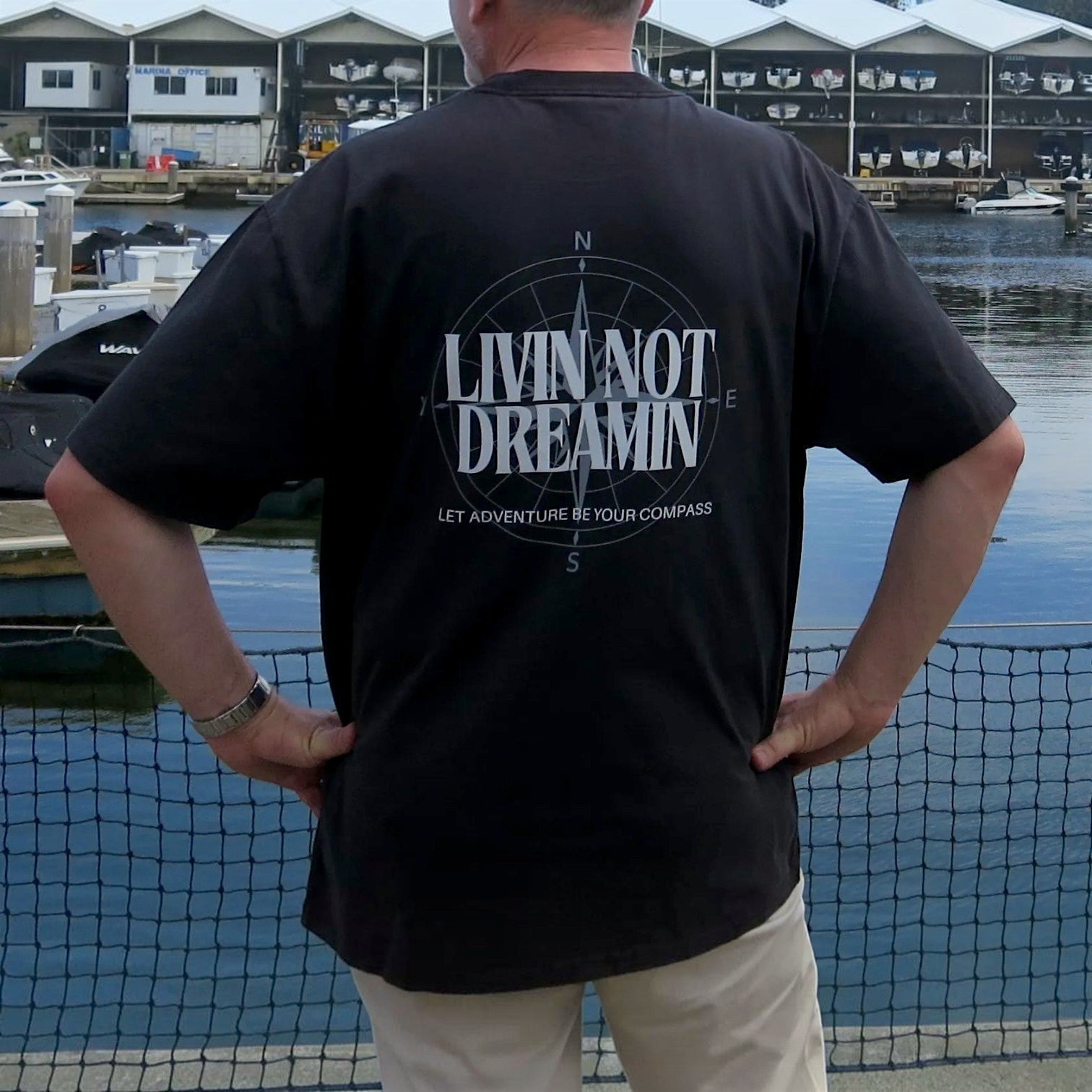 Livin’ not Dreamin’ black heavyweight cotton crew neck t-shirt with a white compass back graphic at a marina
