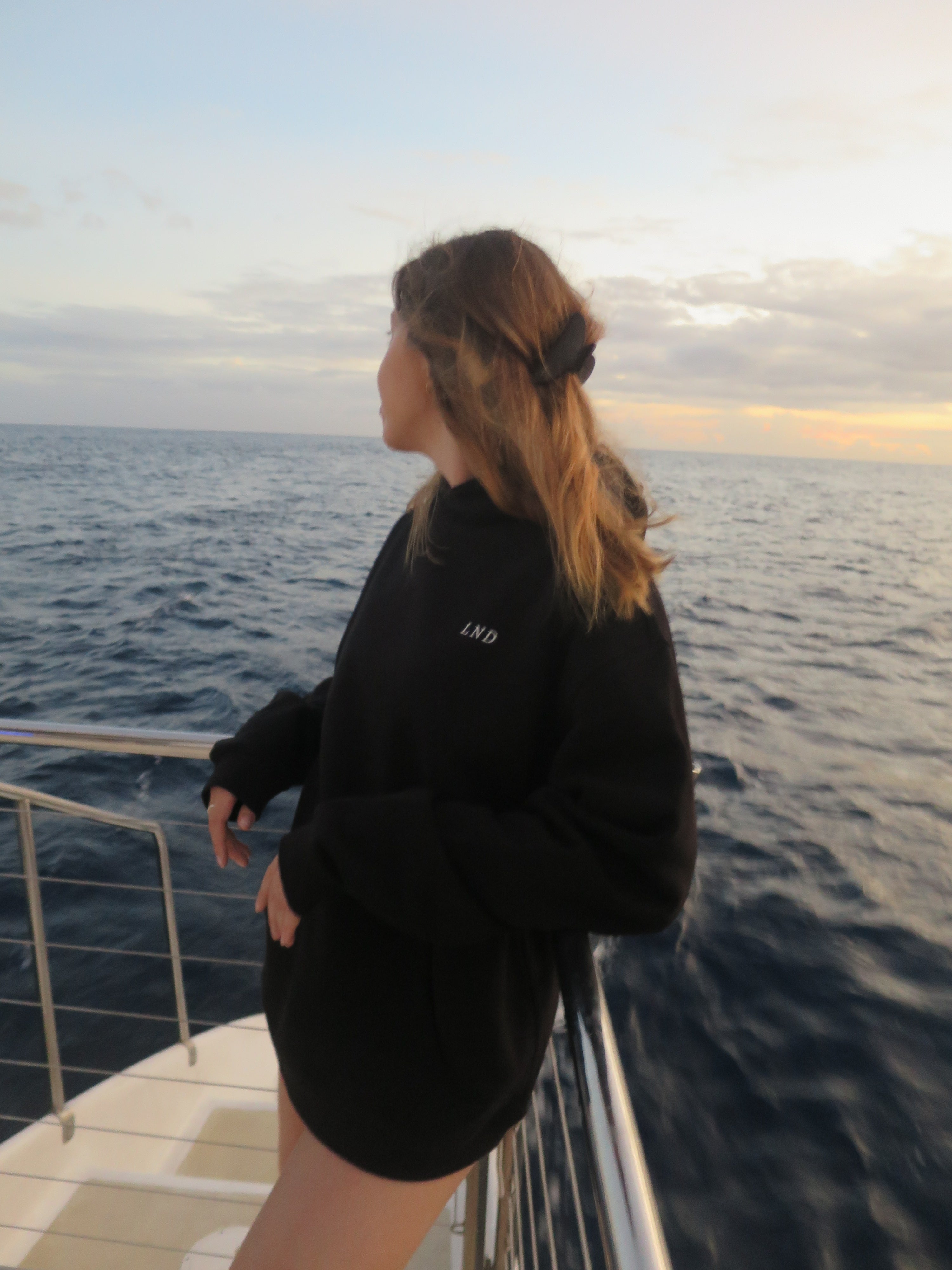 Woman wearing a black Livin’ not Dreamin’ hoodie aboard a boat at sunset