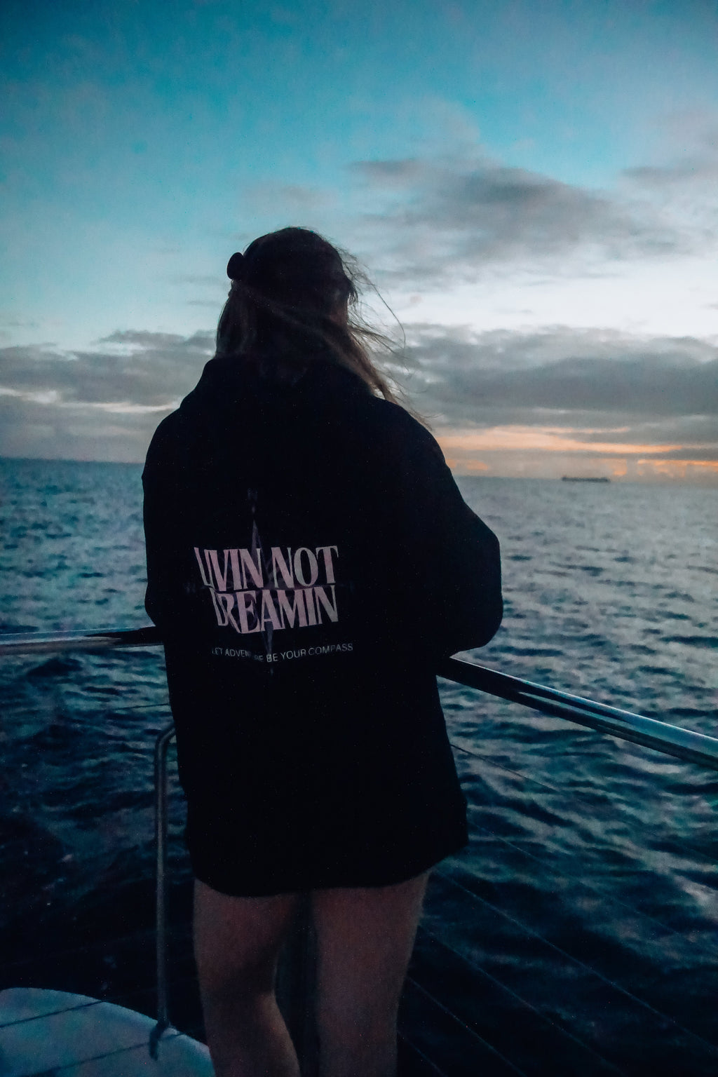 Livin’ not Dreamin’ black fleece-lined hoodie with white logo, worn by a person overlooking the ocean at sunset.