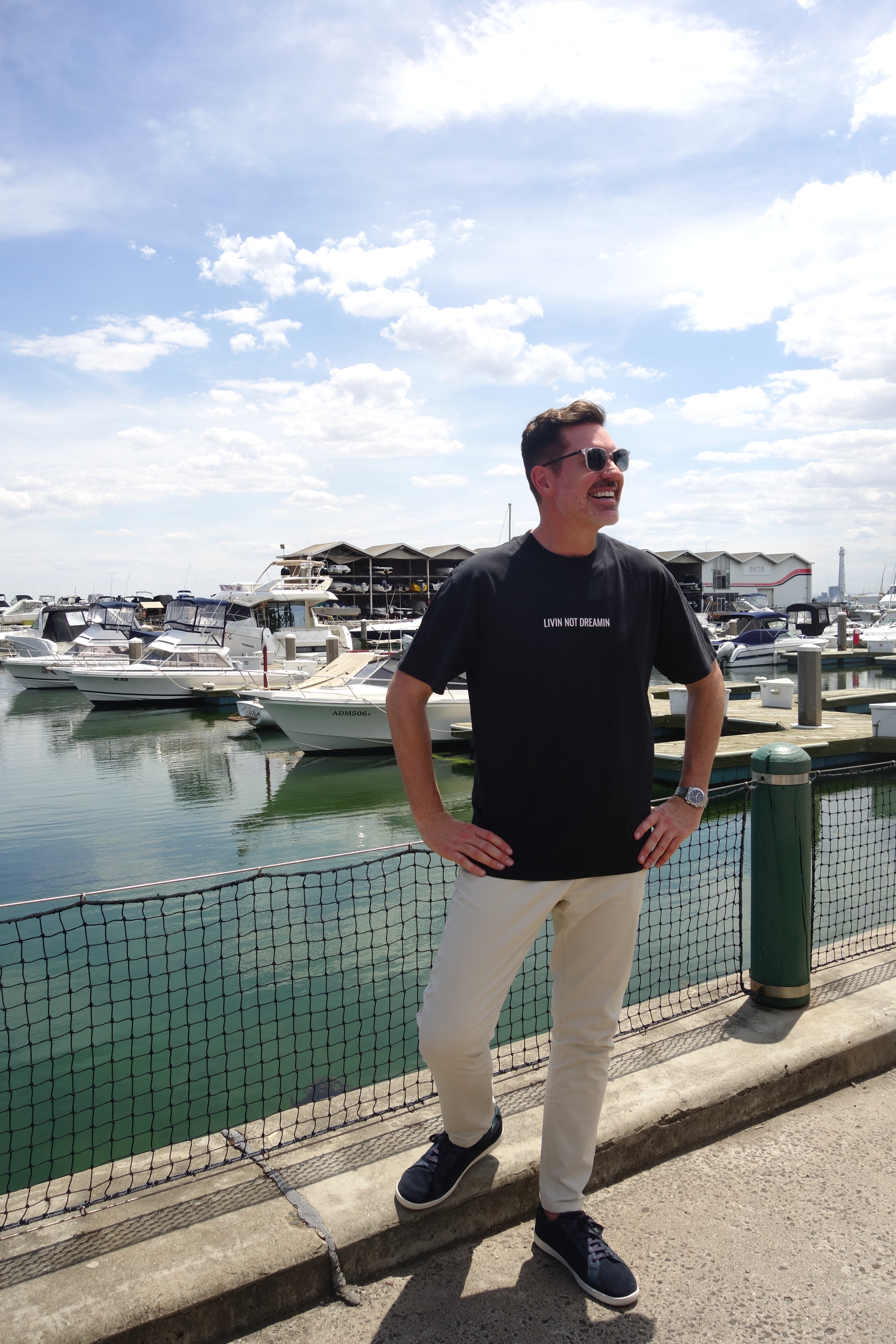 Man wearing a charcoal Livin’ not Dreamin’ Classic T-shirt beside boats at a marina.