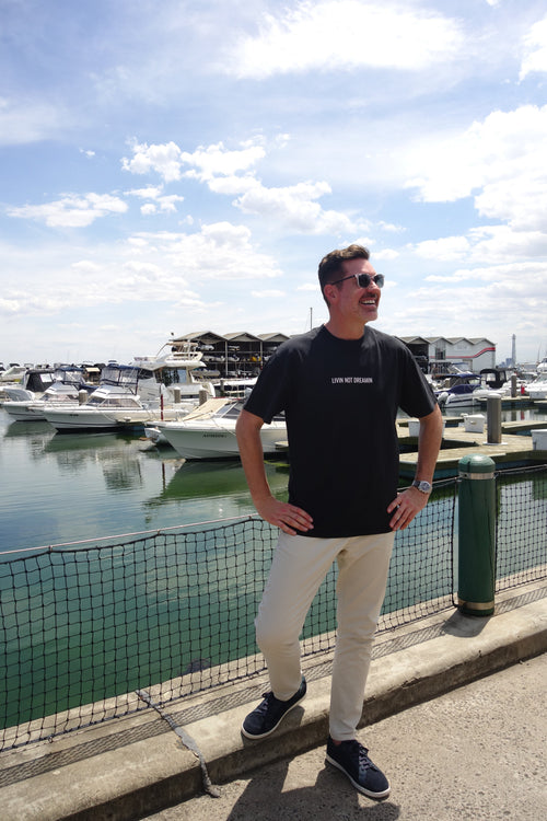 Man wearing a charcoal Livin’ not Dreamin’ Classic T-shirt beside boats at a marina.