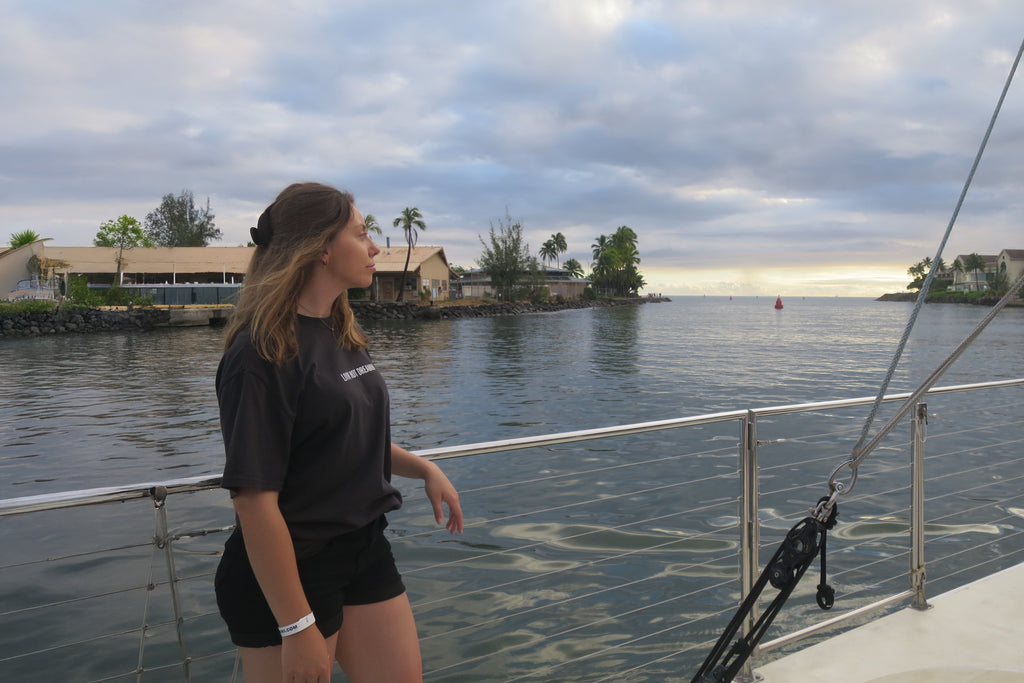 Woman wearing the charcoal black Classic Livin’ not Dreamin’ cotton t-shirt on a boat deck overlooking a coastal harbour.