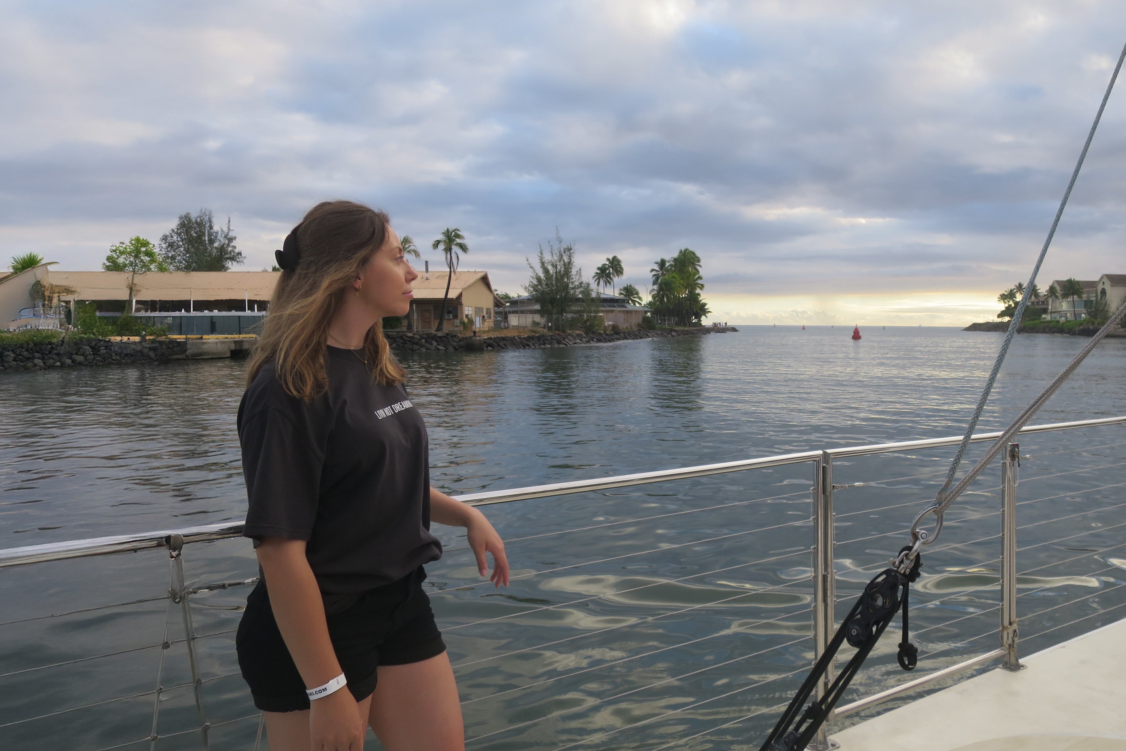 Woman wearing the charcoal black Classic Livin’ not Dreamin’ cotton t-shirt on a boat deck overlooking a coastal harbour.